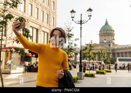 Der junge Mann jemand warten und selfie, er kleidete sich in einen gelben Pullover, einen schwarzen Regenmantel oder Jacke, Jeans, Street und Kazanskiy Kathedrale Stockfoto