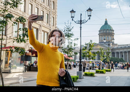 Der junge Mann jemand warten und selfie, er kleidete sich in einen gelben Pullover, einen schwarzen Regenmantel oder Jacke, Jeans, Street und Kazanskiy Kathedrale Stockfoto