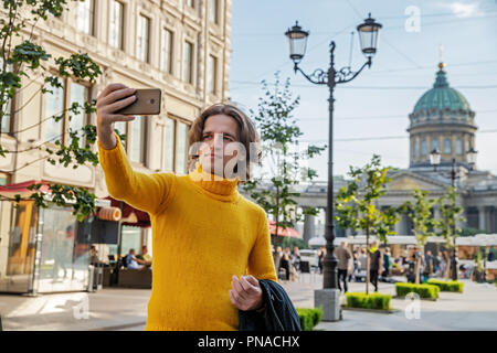 Der junge Mann jemand warten und selfie, er kleidete sich in einen gelben Pullover, einen schwarzen Regenmantel oder Jacke, Jeans, Street und Kazanskiy Kathedrale Stockfoto