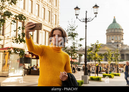 Der junge Mann jemand warten und selfie, er kleidete sich in einen gelben Pullover, einen schwarzen Regenmantel oder Jacke, Jeans, Street und Kazanskiy Kathedrale Stockfoto