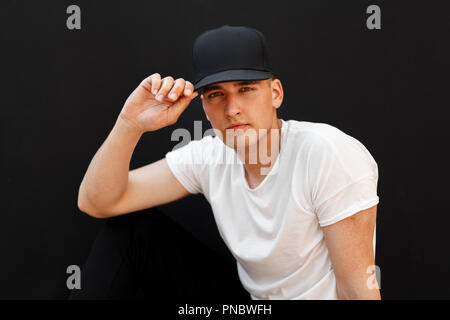 Jungen gutaussehenden Mann in einem modischen schwarzen Kappe und weißen T-Shirt sitzt auf einem schwarzen Hintergrund Stockfoto