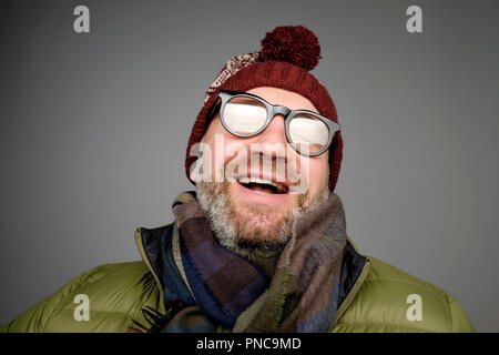 Portrait von Reifen hipster Mann in Gläsern und warme Mütze auf grauem Hintergrund posiert. Lächelnd bärtiger Mann, der warme Kleidung für den Winter Studio gedreht. Clo Stockfoto