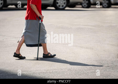 Eine alte Frau mit einem Stock geht über die Straße. Auf Asphalt gesehen Frauen Schatten. Stockfoto