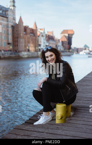 Junge Frau in eine Lederjacke gekleidet sitzt durch den Fluß Pier in Danzig, Polen, Lachen. Stockfoto