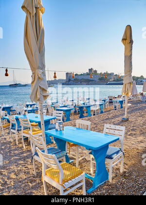 Tische mit Stühlen eines Türkischen Taverne in der Nähe des Meeres in einem Strand der Kumbahce Bucht mit dem Schloss von St. Peter im Hintergrund. Bodrum. Die Türkei. Stockfoto