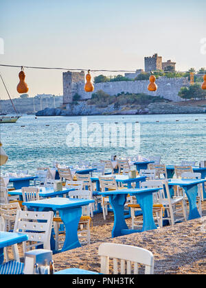 Tische mit Stühlen eines Türkischen Taverne in der Nähe des Meeres in einem Strand der Kumbahce Bucht mit dem Schloss von St. Peter im Hintergrund. Bodrum. Die Türkei. Stockfoto