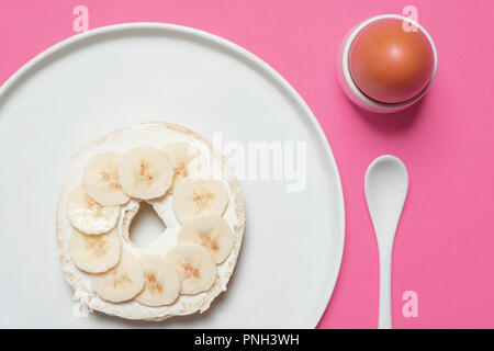 Die Hälfte Bagel mit Frischkäse und ein hart gekochtes Ei auf einem weißen Teller auf einem rosa Hintergrund mit einem weißen Löffel mit Platz für Text Stockfoto