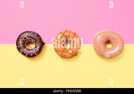 Drei Donuts in einer Reihe auf einem Gelb und Rosa split Hintergrund mit Schokolade besprüht, Normalpapier, Und wirbelte Karamell Stockfoto