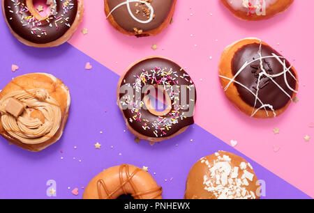 Sortierte Donuts auf einen Split rosa und lila Pastell Hintergrund mit einem klassischen Schokolade Ring und streuseln Donuts in der Mitte. Stockfoto