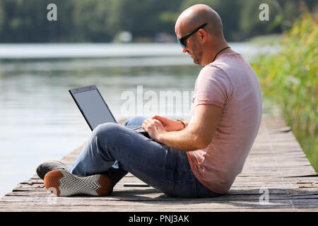 Der 41-jährige sportlich Mann arbeitet in Brandenburg in der Nähe von einem See mit Smartphone und Laptop. Stockfoto