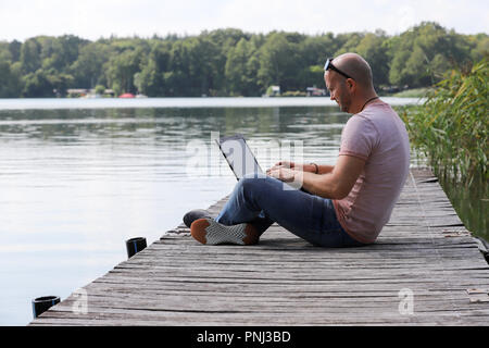 Der 41-jährige sportlich Mann arbeitet in Brandenburg in der Nähe von einem See mit Smartphone und Laptop. Stockfoto