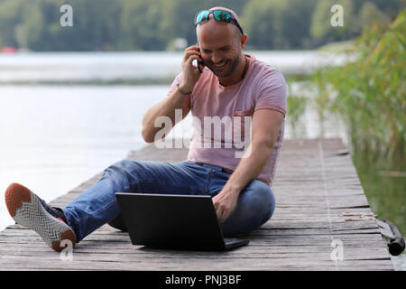 Der 41-jährige sportlich Mann arbeitet in Brandenburg in der Nähe von einem See mit Smartphone und Laptop. Stockfoto
