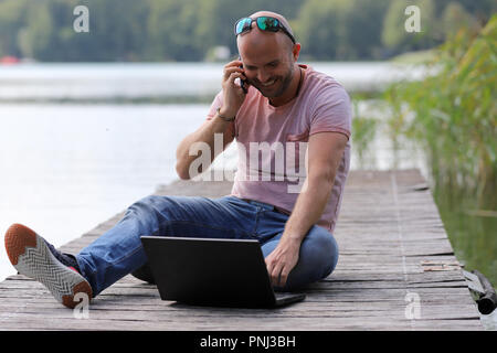 Der 41-jährige sportlich Mann arbeitet in Brandenburg in der Nähe von einem See mit Smartphone und Laptop. Stockfoto