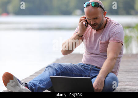 Der 41-jährige sportlich Mann arbeitet in Brandenburg in der Nähe von einem See mit Smartphone und Laptop. Stockfoto