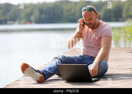 Der 41-jährige sportlich Mann arbeitet in Brandenburg in der Nähe von einem See mit Smartphone und Laptop. Stockfoto