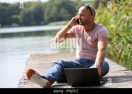 Der 41-jährige sportlich Mann arbeitet in Brandenburg in der Nähe von einem See mit Smartphone und Laptop. Stockfoto