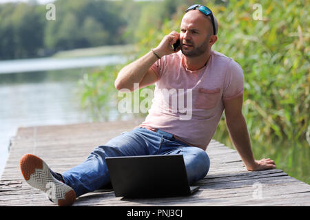 Der 41-jährige sportlich Mann arbeitet in Brandenburg in der Nähe von einem See mit Smartphone und Laptop. Stockfoto