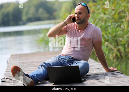 Der 41-jährige sportlich Mann arbeitet in Brandenburg in der Nähe von einem See mit Smartphone und Laptop. Stockfoto