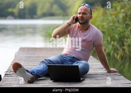 Der 41-jährige sportlich Mann arbeitet in Brandenburg in der Nähe von einem See mit Smartphone und Laptop. Stockfoto