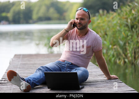 Der 41-jährige sportlich Mann arbeitet in Brandenburg in der Nähe von einem See mit Smartphone und Laptop. Stockfoto