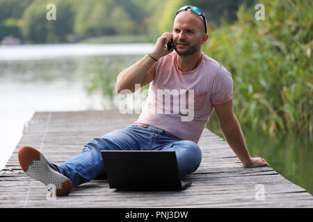 Der 41-jährige sportlich Mann arbeitet in Brandenburg in der Nähe von einem See mit Smartphone und Laptop. Stockfoto