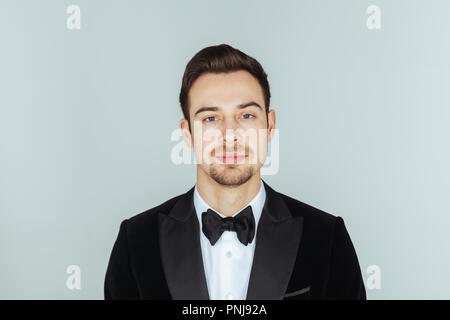 Portrait einer jungen gutaussehenden Mann im Smoking, in die Kamera schaut, gegen Pl​ain studio Hintergrund. Stockfoto