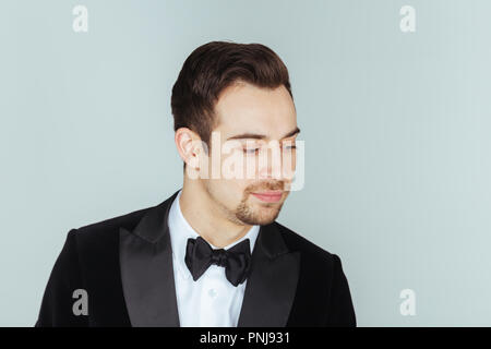 Portrait einer jungen gutaussehenden Mann im Smoking, lächelnd und mit Blick auf die Kamera, gegen Pl​ain studio Hintergrund. Stockfoto