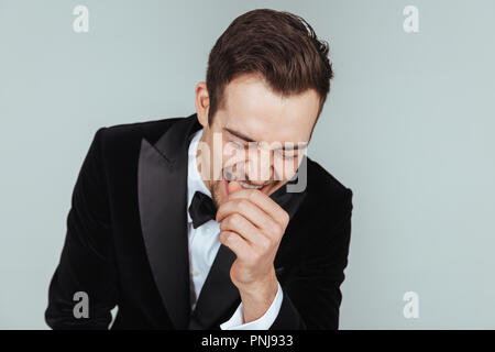 Portrait einer jungen gutaussehenden Mann im Smoking, Lachen, die Hand in der Nähe von Gesicht, gegen Pl​ain studio Hintergrund. Stockfoto