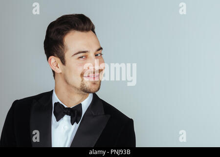 Portrait einer jungen gutaussehenden Mann im Smoking, lächelnd und mit Blick auf die Kamera, gegen Pl​ain studio Hintergrund. Stockfoto