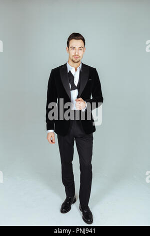 Portrait einer jungen gutaussehenden Mann in Anzug, stehend und mit Blick auf die Kamera, Bow Tie rückgängig gemacht, gegen normales studio Hintergrund Stockfoto