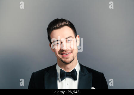 Portrait einer jungen gutaussehenden Mann im Smoking, lächelnd und mit Blick auf die Kamera, gegen normales studio Hintergrund Stockfoto