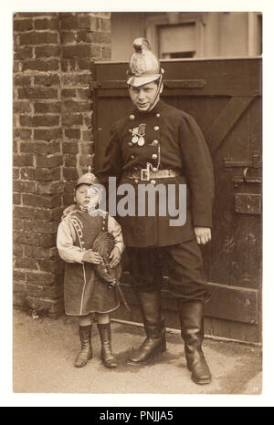 Originalfotopostkarte eines Feuerwehrhelden in Uniform mit Medaillen, die draußen für ein Foto mit seinem jungen Sohn posieren, der sich als Feuerwehrmann mit Feuerwehruniform verkleidet und einen Feuerwehrschlauch hält, um 1910, Manchester, Großbritannien Stockfoto