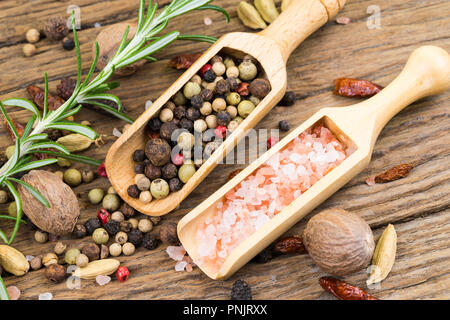 Nahaufnahme der bunte Pfefferkörner und rosa Kristall Salz auf kleinen hölzernen Spice Lufthutzen auf rustikalen Holz Hintergrund mit einem Zweig Rosmarin in der backgroun Stockfoto