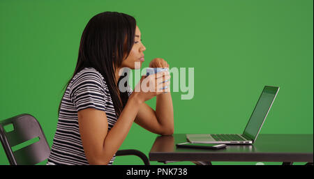Profil von schwarzes Weibchen sitzen, Kaffee trinken und mit Laptop auf dem Green Screen Stockfoto