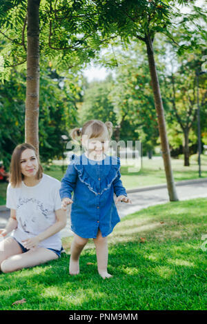 Portrait von 3 Jahre alten Mädchen von ihrer Mutter beim Spielen auf einem Rasen in einem Stadtpark an einem warmen sonnigen Tag Stockfoto