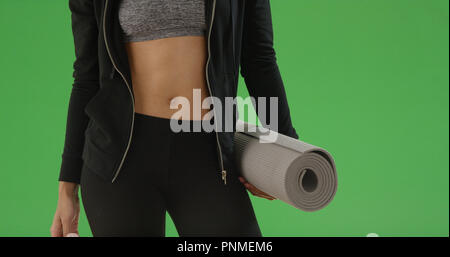 Junge schwarze Frau steht in ihrem Crop Top mit Yoga Matte auf Green Screen Stockfoto