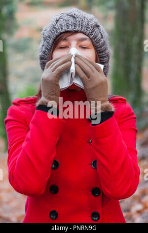 Junge Frau mit Erkältung oder Grippe blasen Nase, Niesen auf weißem Papier Taschentuch in Wald trägt einen roten Mantel oder Jacke, eine Mütze ein Stockfoto