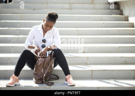 Afrikanische Frau mit smart phone sitzend auf weißen Treppe im Freien. Stockfoto