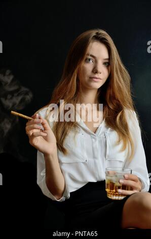 Junge Weibchen sitzt mit Zigarillo und Glas Whisky in der Hand. Elegante Frau mit weißer Spitze und schwarzen Kleid. Stockfoto