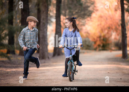 Mädchen und Jungen auf einem Fahrrad neben laufen auf dem Weg zu einem Park Stockfoto