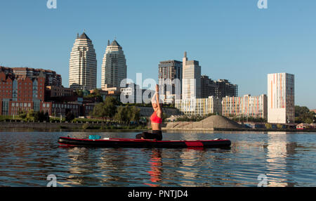 Stand Up Paddle Board Yoga von schönen Mädchen auf der helle Stadt Hintergrund durchgeführt, Yoga Ausbildung auf dem Wasser Stockfoto