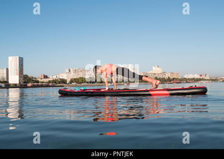 Stand Up Paddle Board Yoga von schönen Mädchen auf der helle Stadt Hintergrund durchgeführt, Yoga Ausbildung auf dem Wasser Stockfoto