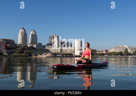 Stand Up Paddle Board Yoga von schönen Mädchen auf der helle Stadt Hintergrund durchgeführt, Yoga Ausbildung auf dem Wasser Stockfoto