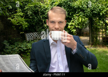 Business Mann in Anzug mit offenem Hemd mit Tasse Kaffee am Frühstückstisch im Garten beim Lesen der Zeitung Stockfoto