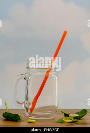 Ein leeres Glas Schale mit einem orangefarbenen Stroh von der Seite gesehen, auf Holztisch, serviert mit Nahrungsergänzungsmitteln, gegen bewölkter Himmel Hintergrund und viel Stockfoto