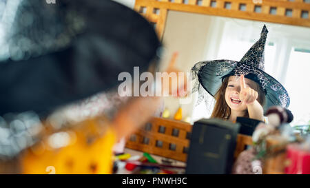 Adorable junge Mädchen mit Hexenhut mit Blick auf sich selbst im Spiegel und zeigt mit dem Finger und Lachen. Glückliches Kind in Halloween Kostüm in suchen Stockfoto