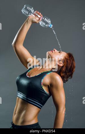 Nach muskulös athletisch Fitness für Frauen im Sport Kleidung gießt Wasser aus der Flasche nach dem Training. Grauer Hintergrund, rothaarige Frau. Stockfoto