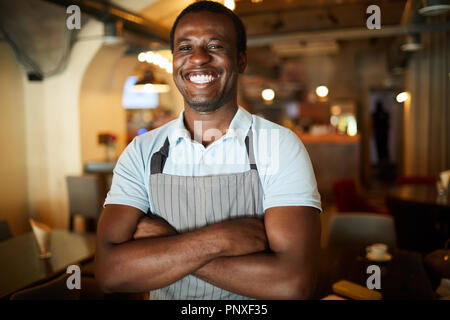 Junge lachen Kellner mit wite toothy Lächeln verschränkte seine Arme auf der Brust im Cafe Stockfoto