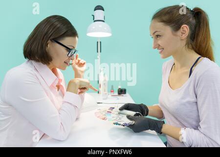 Junge Mädchen Kosmetikerin Maniküre spricht mit einem weiblichen Client, Maniküre, wählt die Farbe von Nagellack, kommuniziert und lächelt. Stockfoto