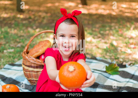 Kleine Mädchen im roten Kleid, mit einem Kürbis, lächelnd. Herbst Picknick im Park auf der Plaid Warenkorb. Halloween Stockfoto